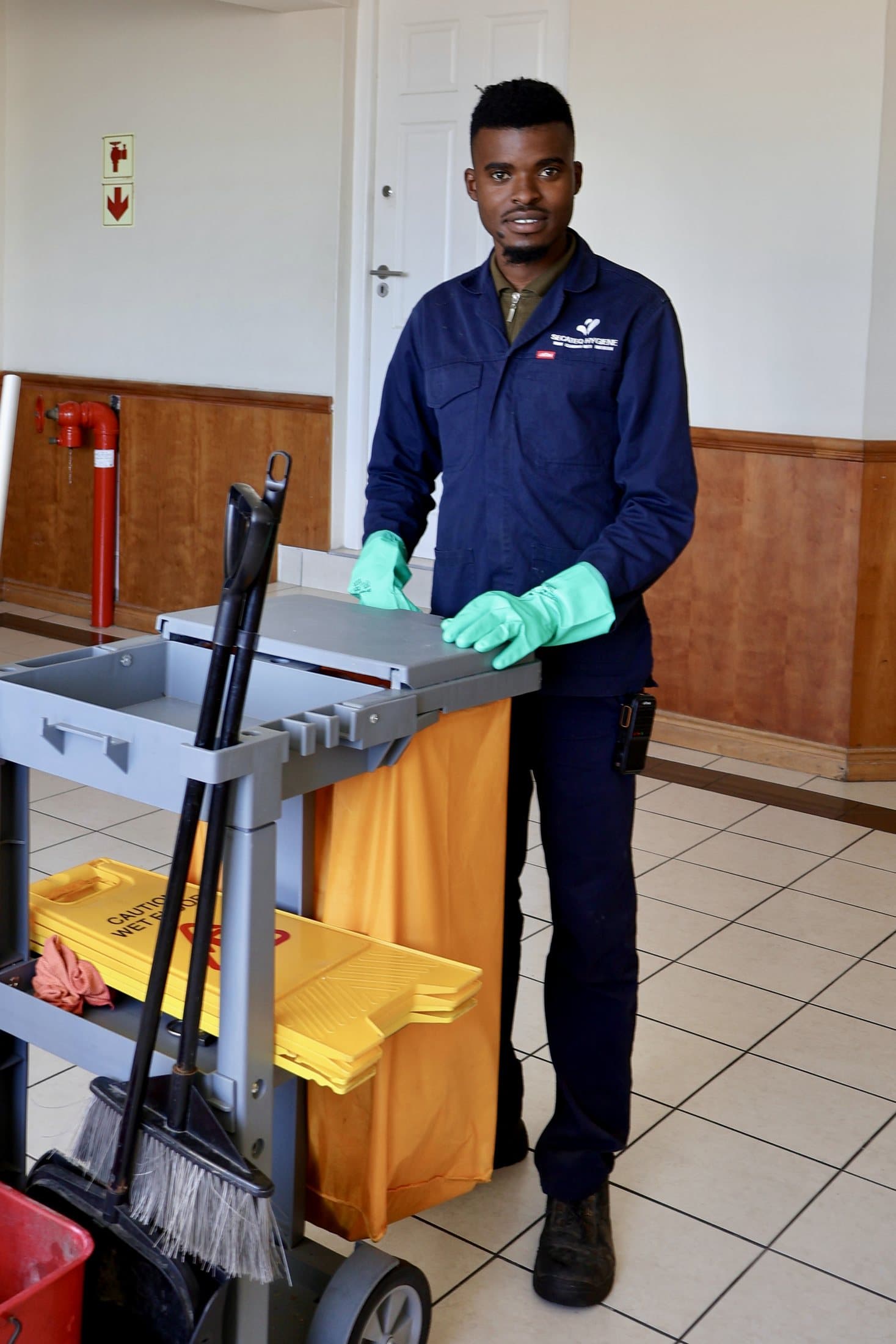 A SecaTeq Hygiene team member in the division's navy overall and green gloves with a fully stocked cleaning trolley in a lift lobby